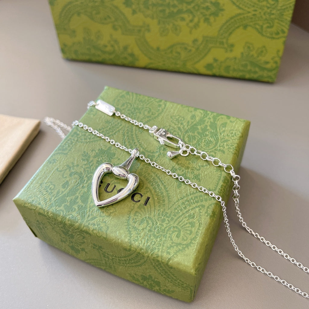 Silver charm necklace with heart pendant and tag accents displayed on green box