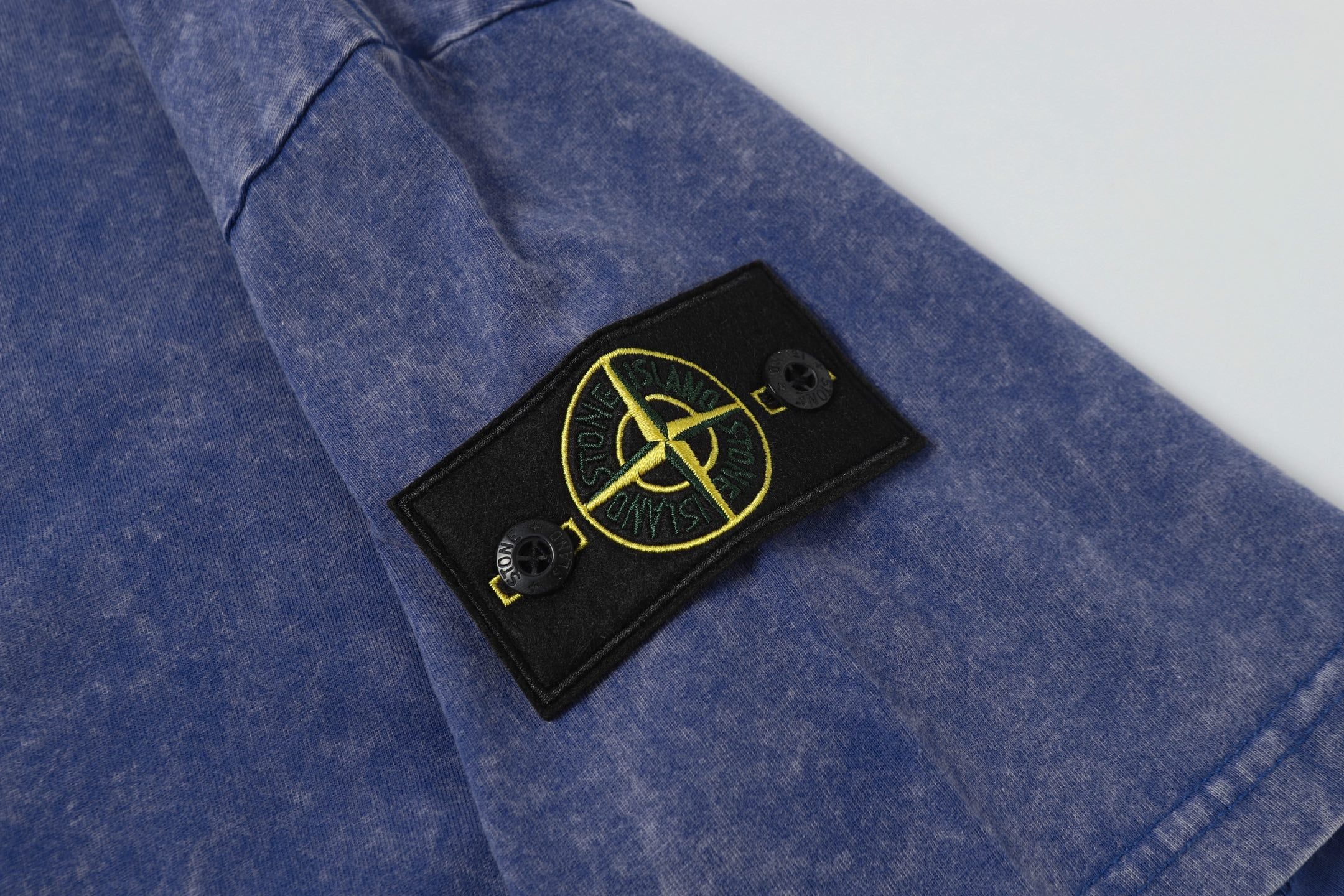 Blue T-shirt with black logo patch and yellow emblem near chest.