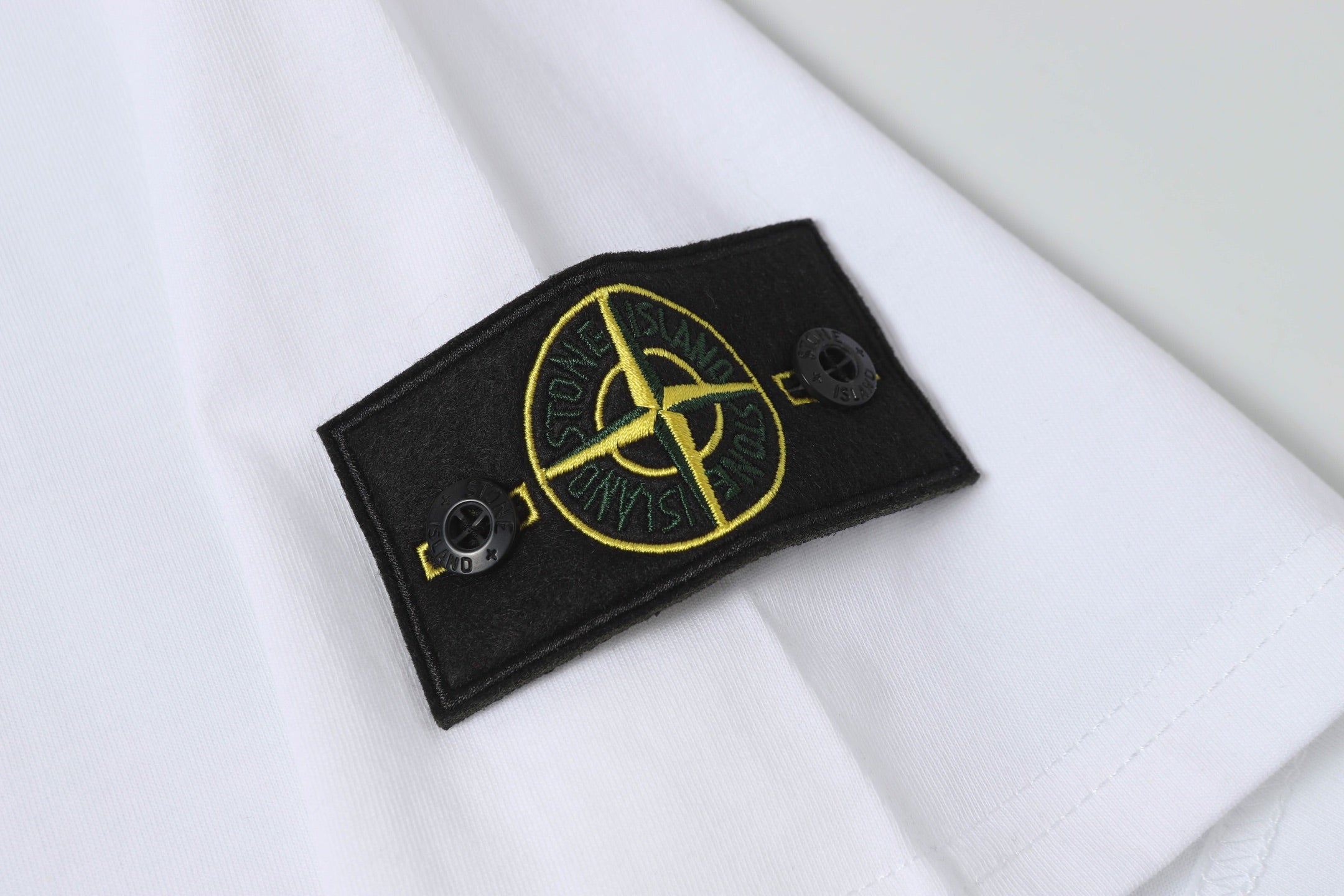 White Stone Island T-shirt sleeve patch with gold emblem on black fabric.
