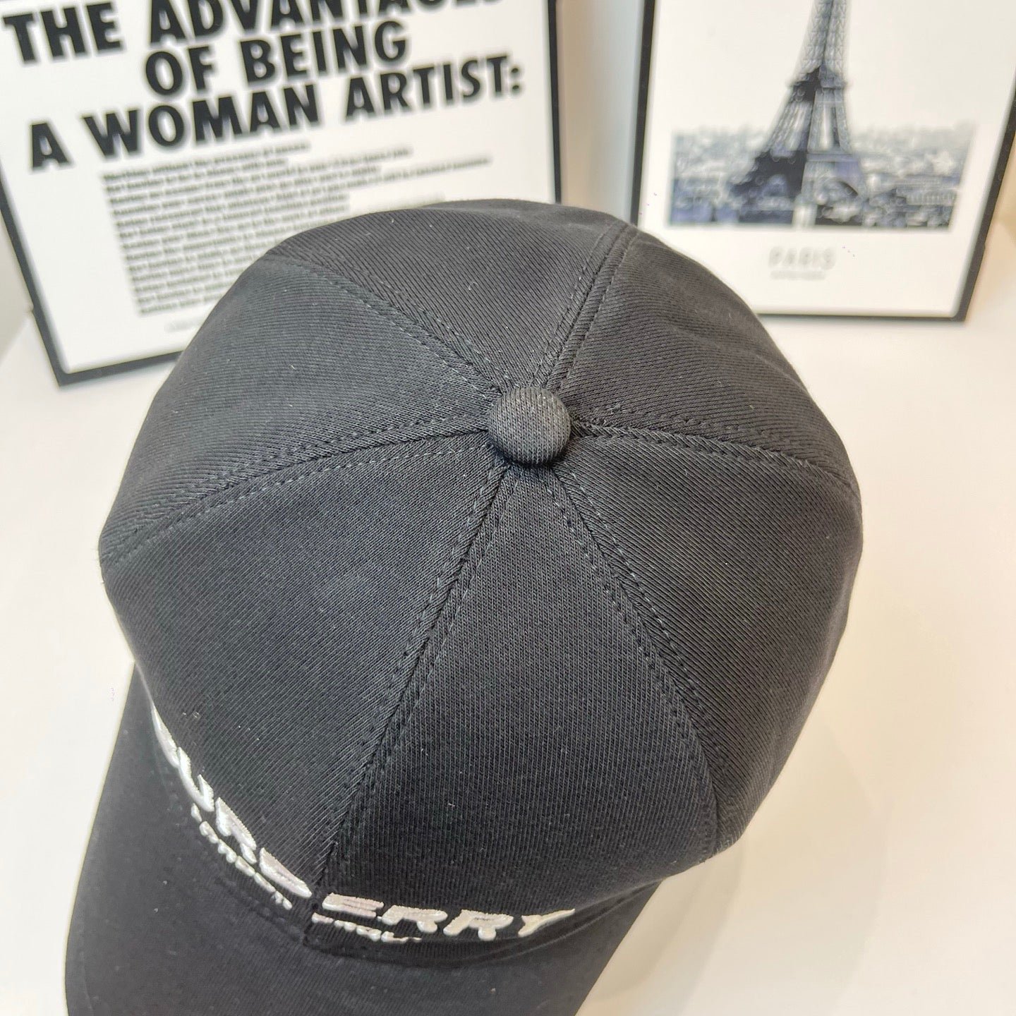 Black Burberry cap showing top panel seam and domed button; side logo partially visible.