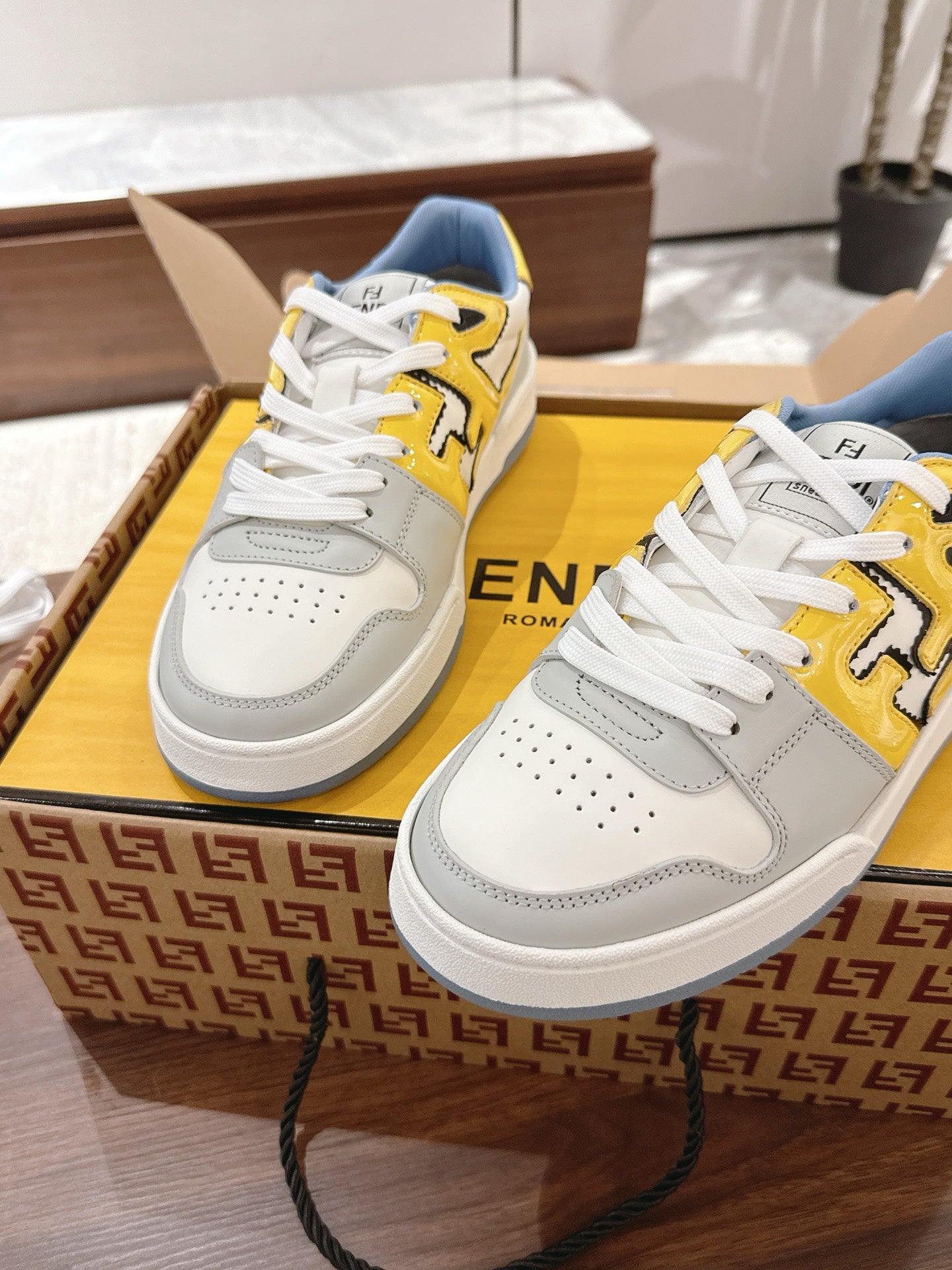 Low-top sneakers, white with yellow logo accents, blue collar, on Fendi box