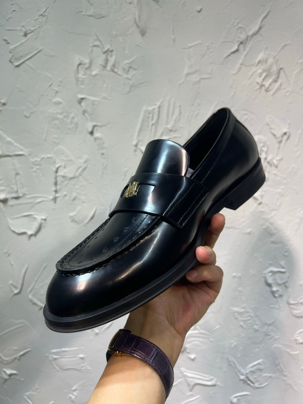 Amiri black leather loafer with gold emblem on strap, held shoe side view