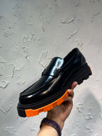 Black leather loafer with orange tread sole, close-up showing metal buckle detail