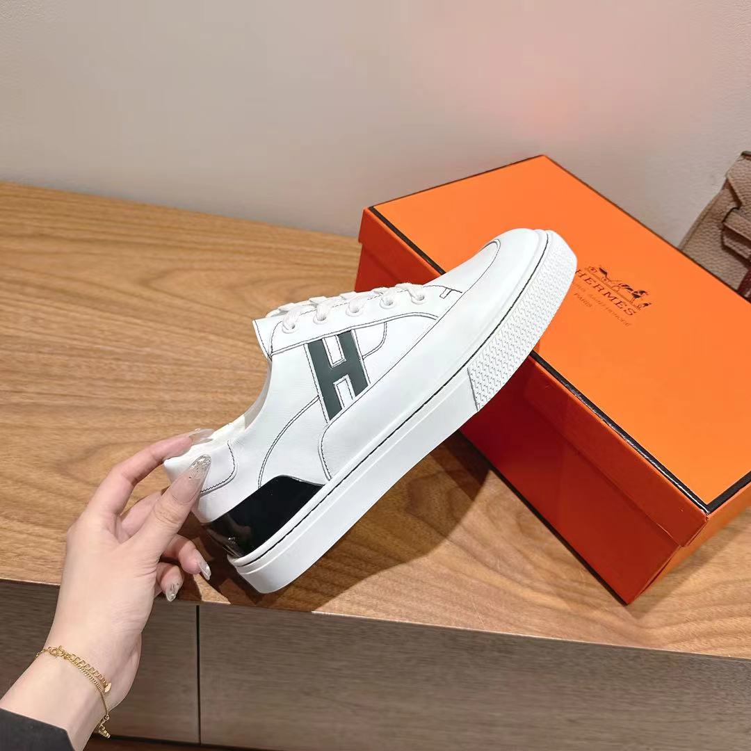 White Hermes unisex sneakers with black sole trim, held near orange shoe box.