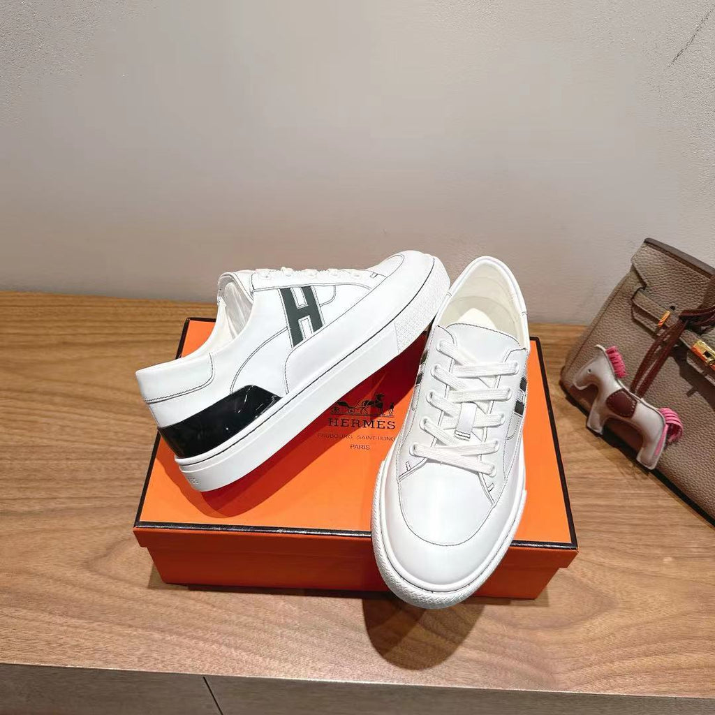 White Hermes low-top sneakers with black side panel and lace-up eyelets, on orange box.
