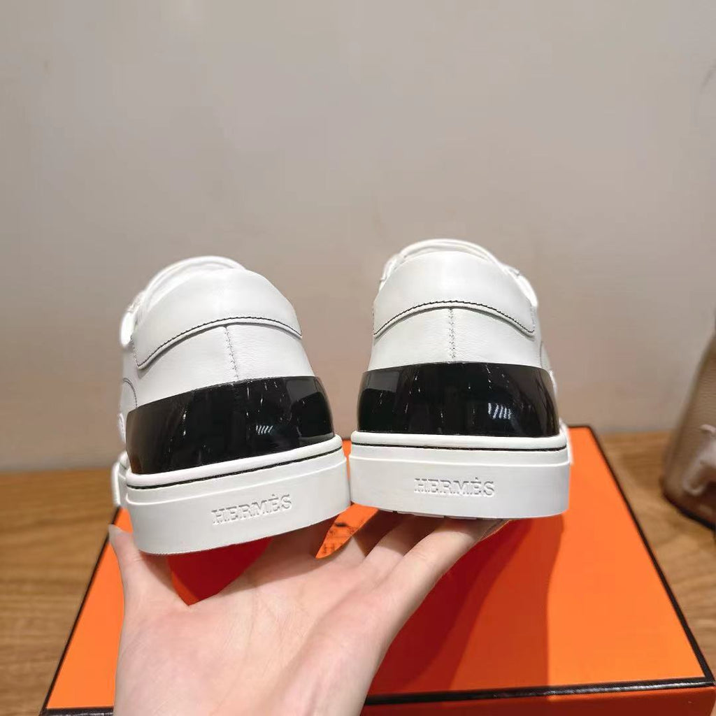 Back view of white Hermes sneakers with black heel panel and logo, on orange box.