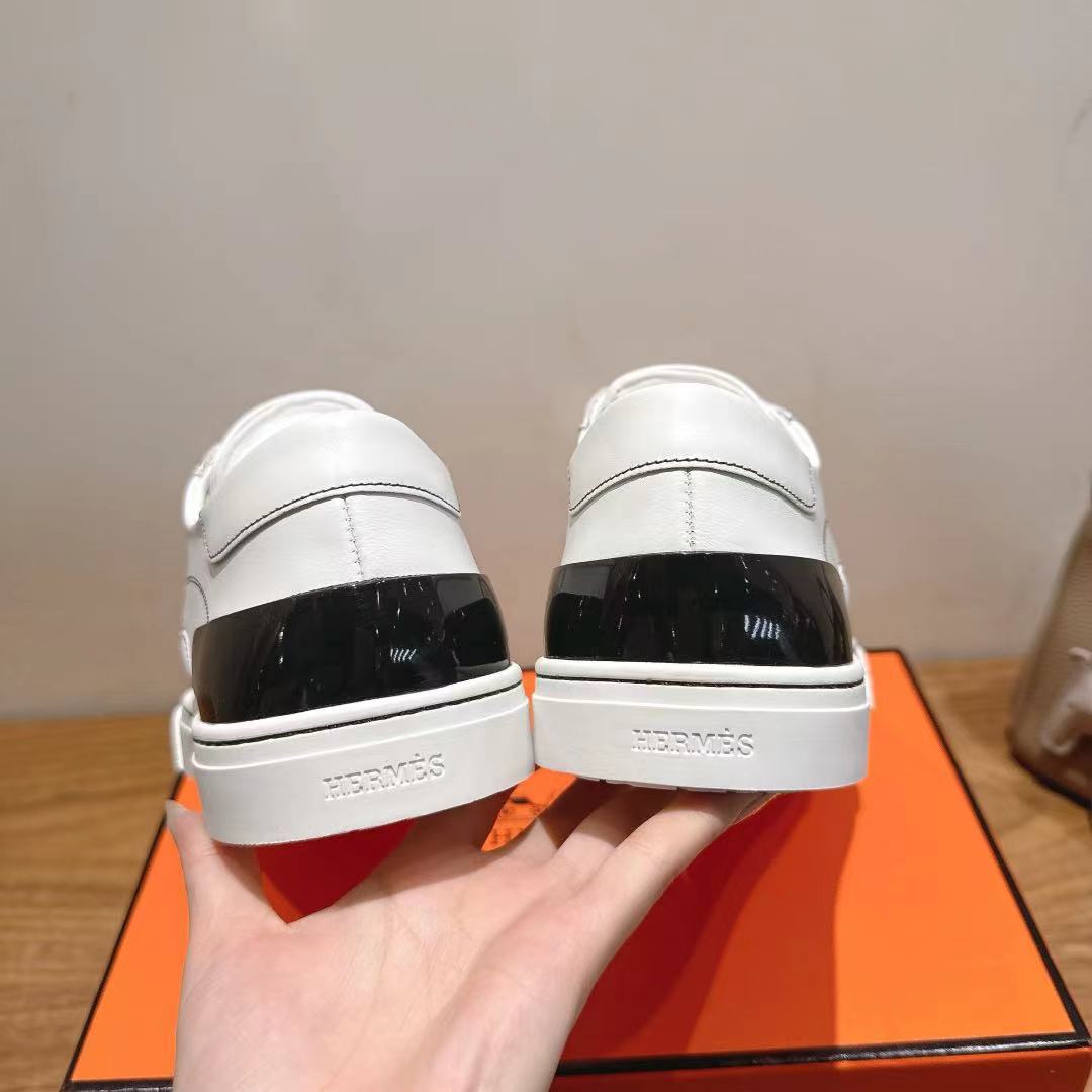 Back view of white Hermes sneakers with black heel panel and logo, on orange box.