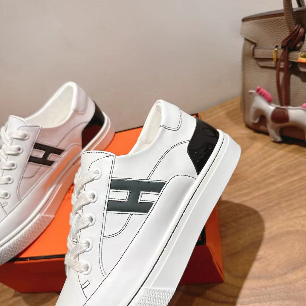 White Hermes unisex sneakers with black logo stripe, close side view on orange box.