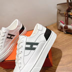 White Hermes unisex sneakers with black logo stripe, close side view on orange box.