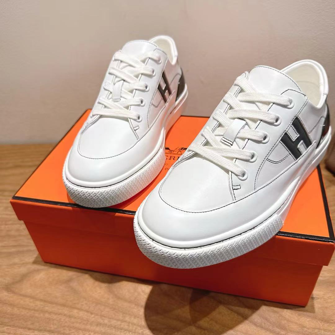 White Hermes unisex luxury shoes with black side H logo, on orange box.