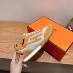 Hermes unisex sneaker close-up on orange box, tan suede, white laces, mesh side.