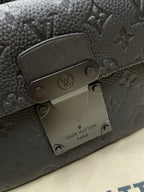 Louis Vuitton men's bag hardware close-up with engraved logo plate and dark monogram texture