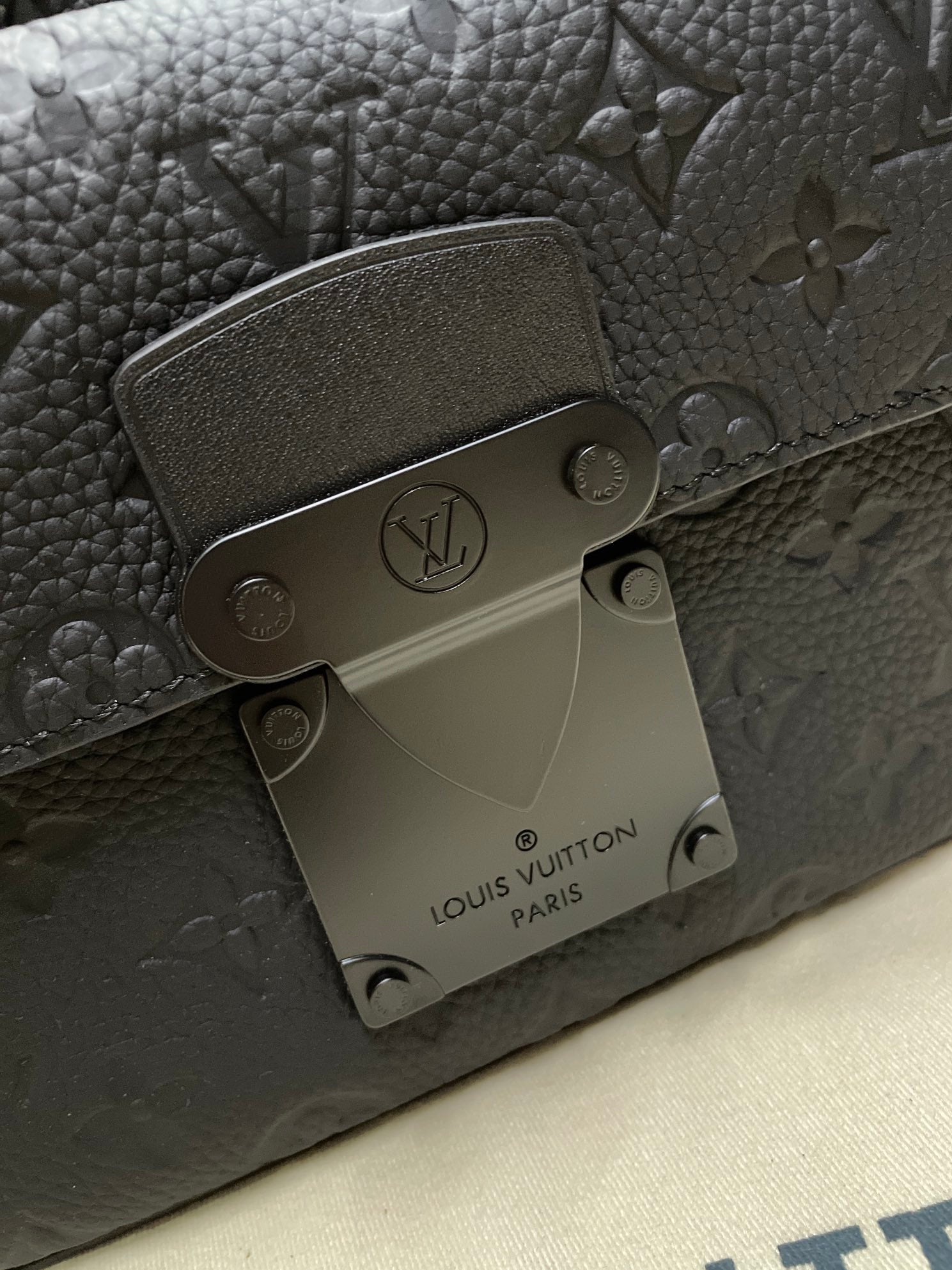 Louis Vuitton men's bag hardware close-up with engraved logo plate and dark monogram texture