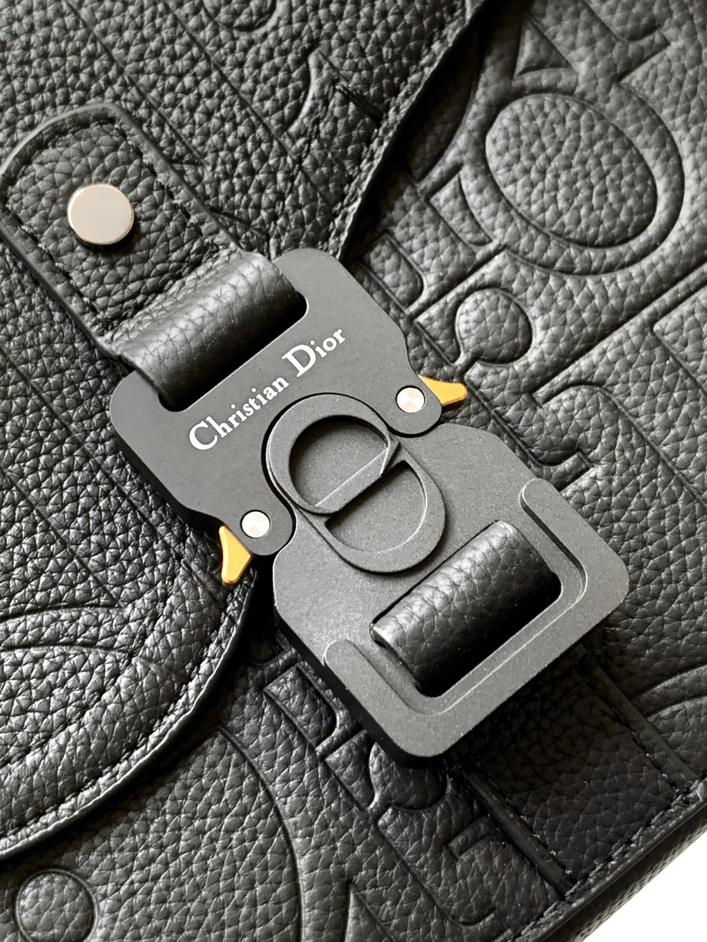 Close-up of Dior metal emblem charm on dark leather flap with belt-style buckle