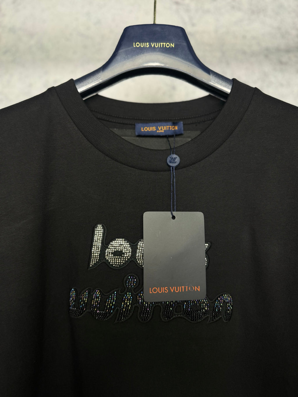 Black T-shirt neckline with rhinestone logo and hanging tag on a hanger