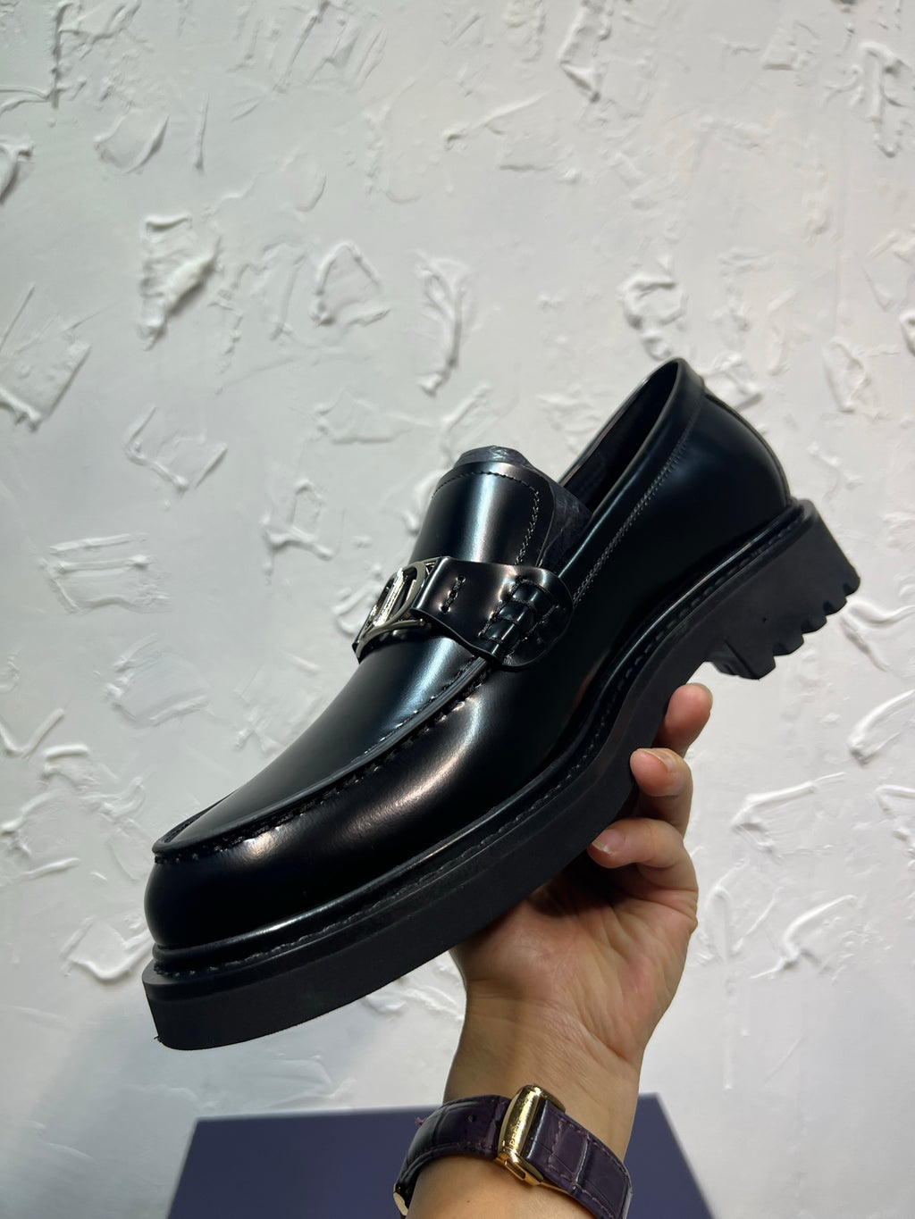 Black leather loafer with square buckle strap and thick sole, held in hand