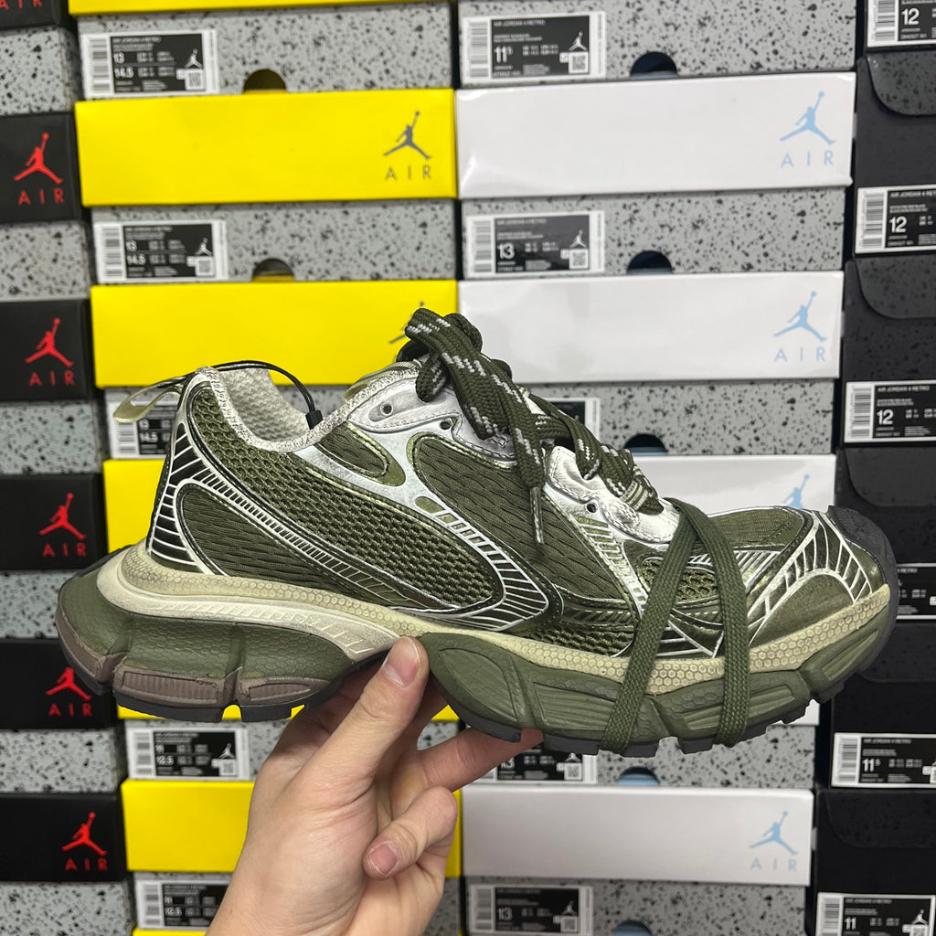 Green and silver sport sneakers with laces, held up against stacked shoe boxes
