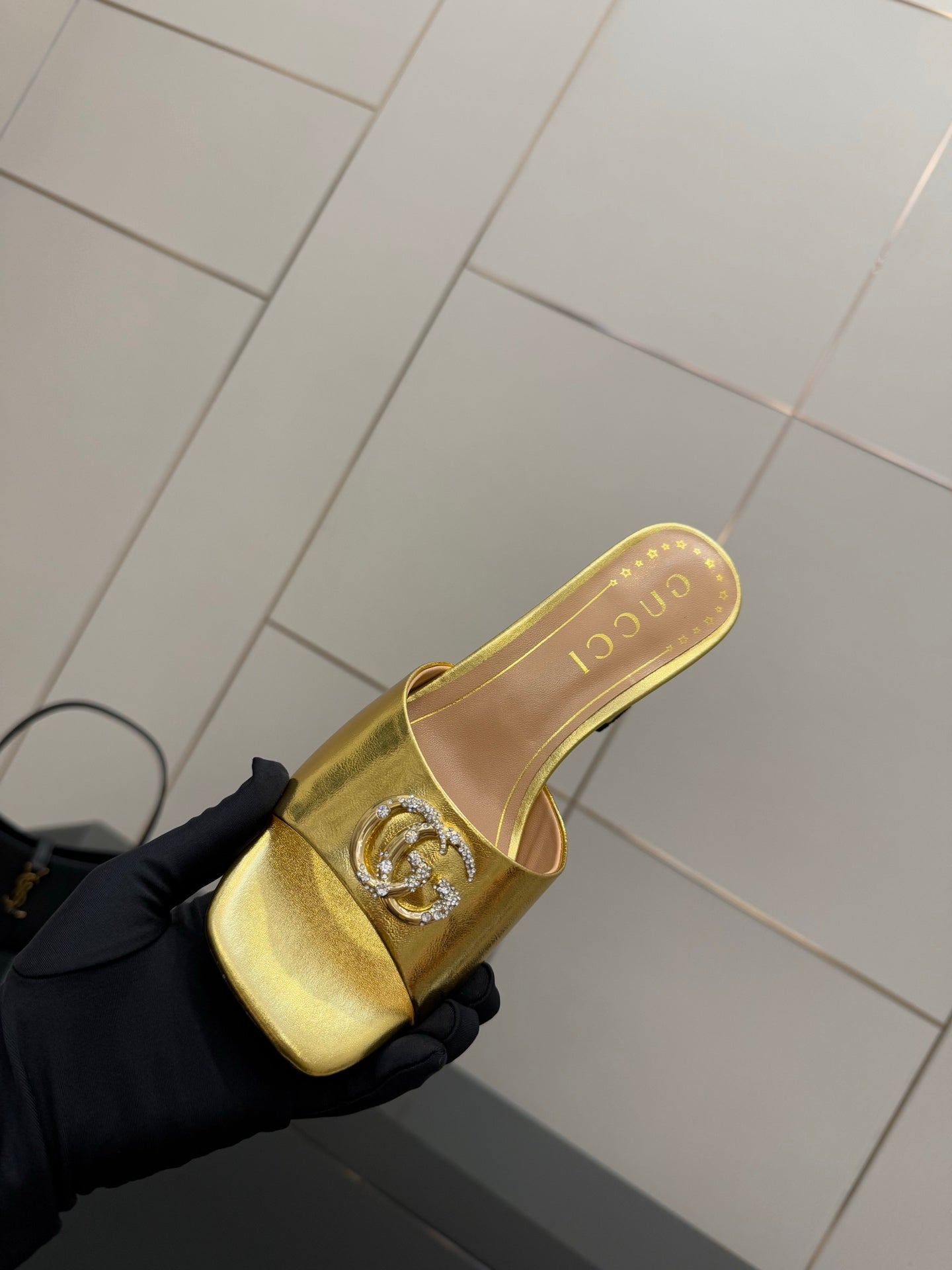 Gucci gold mule on black-gloved hand, showing embossed emblem and open toe