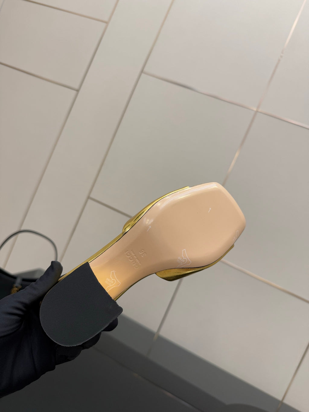 Gucci gold mule sole on black-gloved hand, printed logo on beige footbed