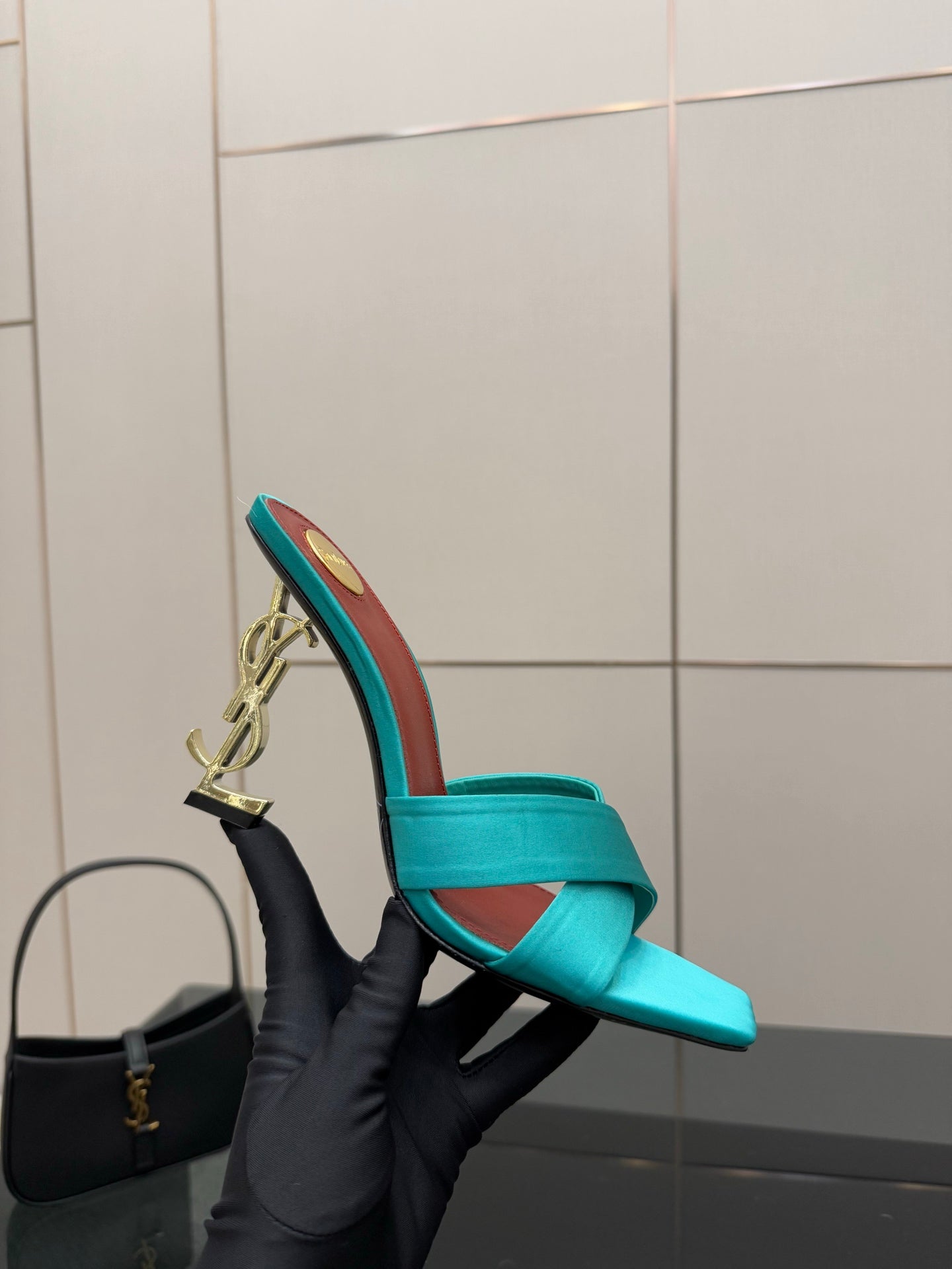 Teal YSL mule sandal held in black glove, open toe with gold logo heel