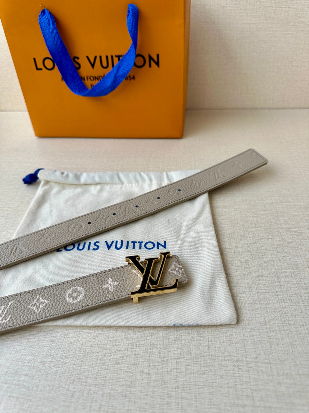 Gray monogram belt with LV buckle on white pouch, blue tie loop visible
