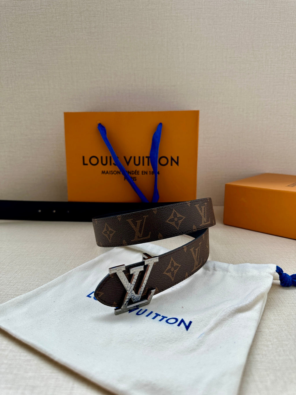 Brown monogram belt with silver LV buckle and blue accents on white dust pouch.