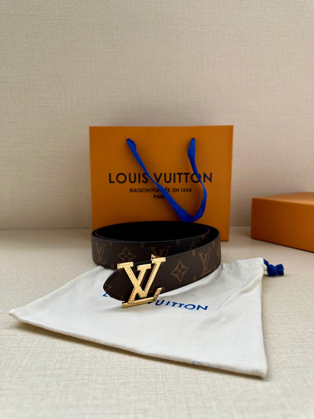Brown monogram belt with silver-tone LV buckle and blue tassel, on white dust bag.