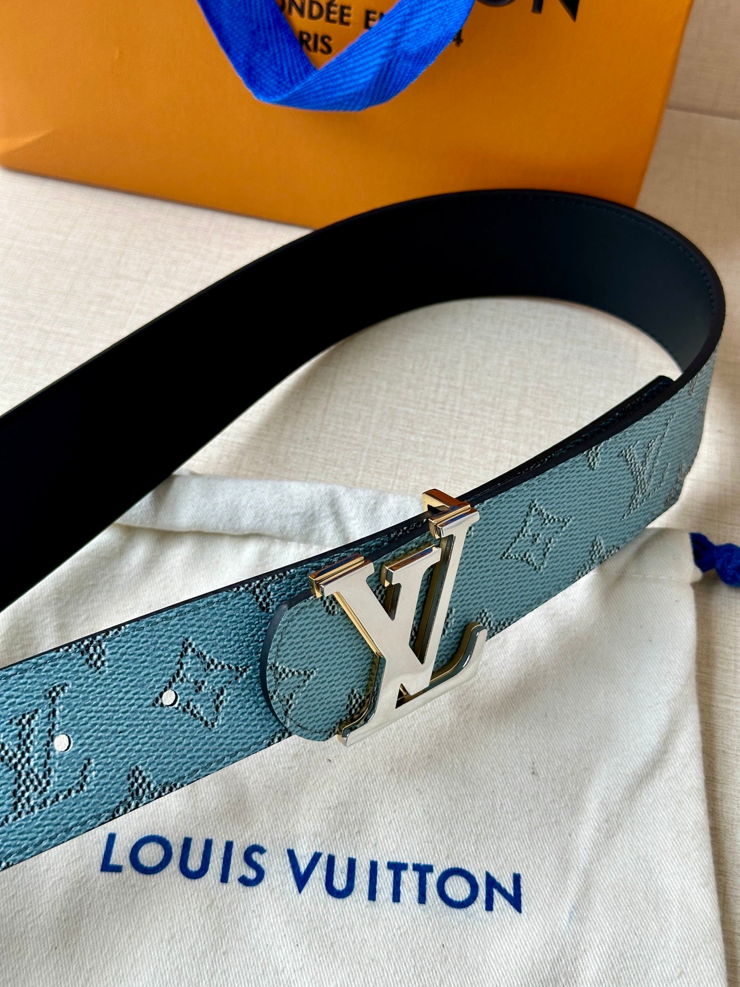 Louis Vuitton belt close-up with blue monogram strap and gold LV buckle on light fabric.