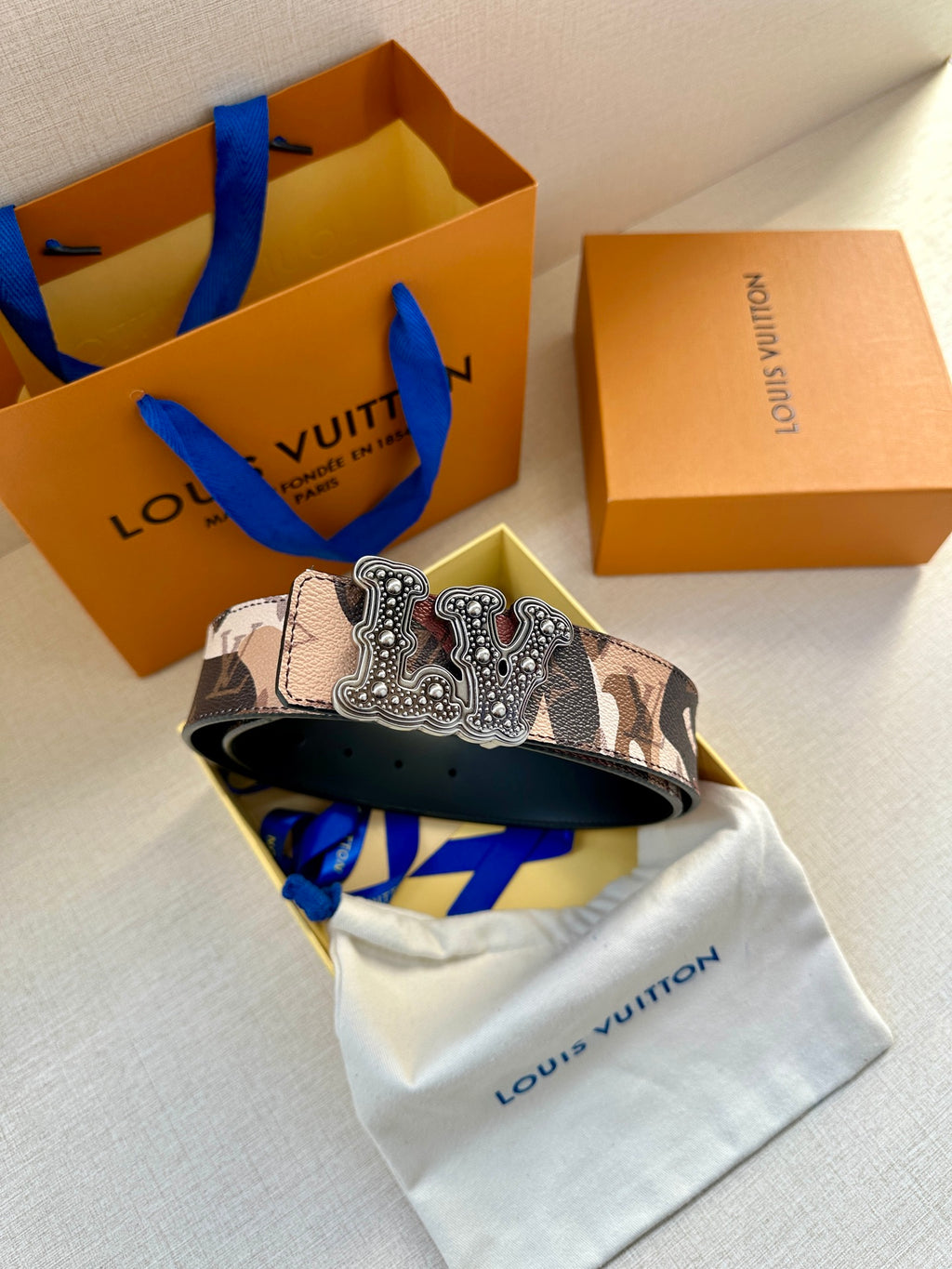 Camo designer belt with wide strap and silver-toned ornate buckle over dust bag