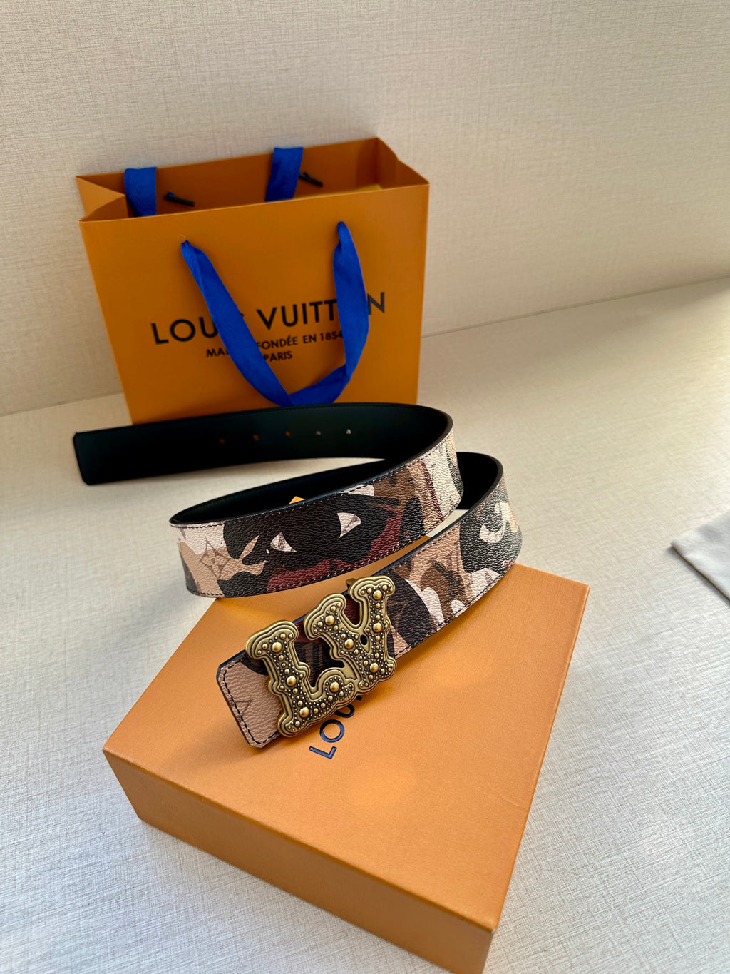 Designer belt on box with brown camo strap and gold-tone decorative buckle