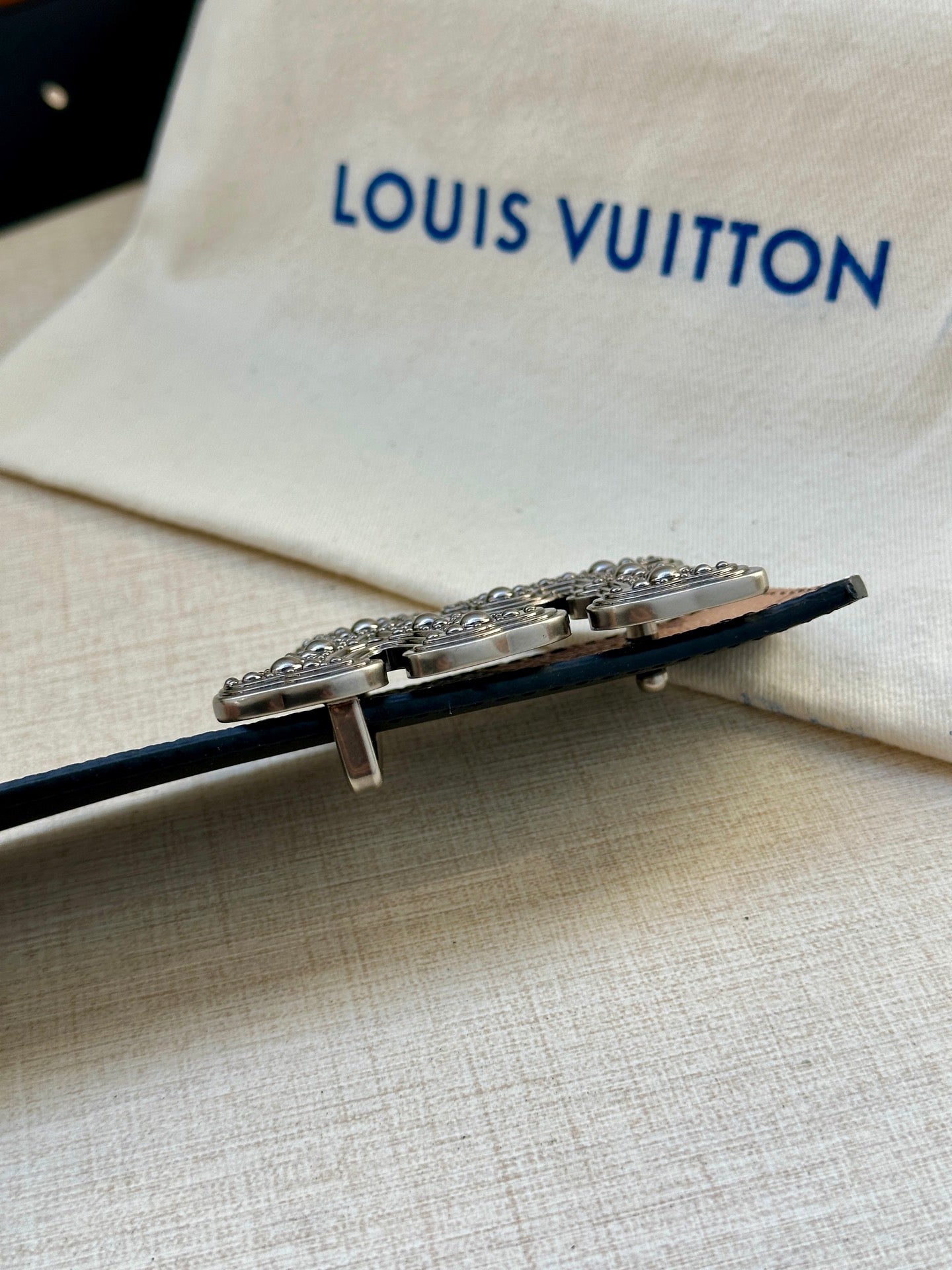 Belt buckle detail with silver-toned hardware on dark strap, resting on dust bag