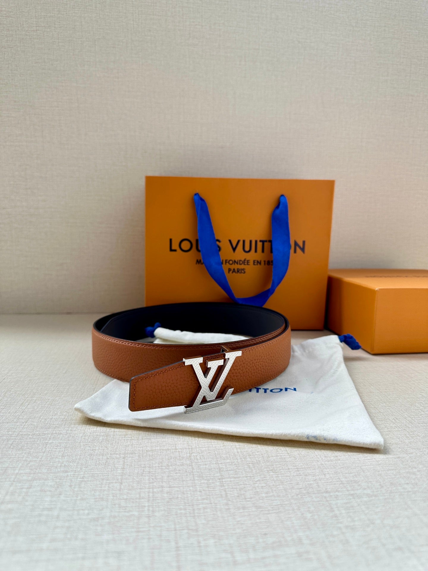Leather belt with brown strap and silver LV buckle, on white dust bag.