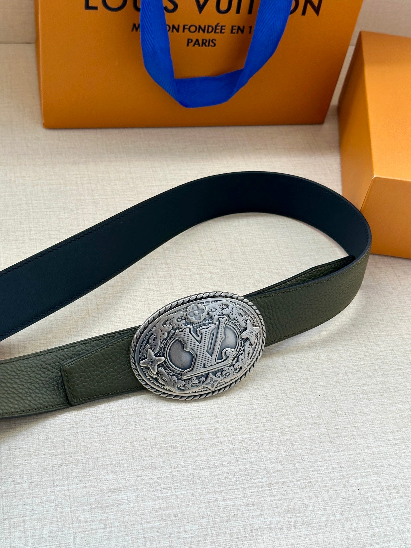 Dark green leather belt with oval embossed logo buckle, angled close-up view.