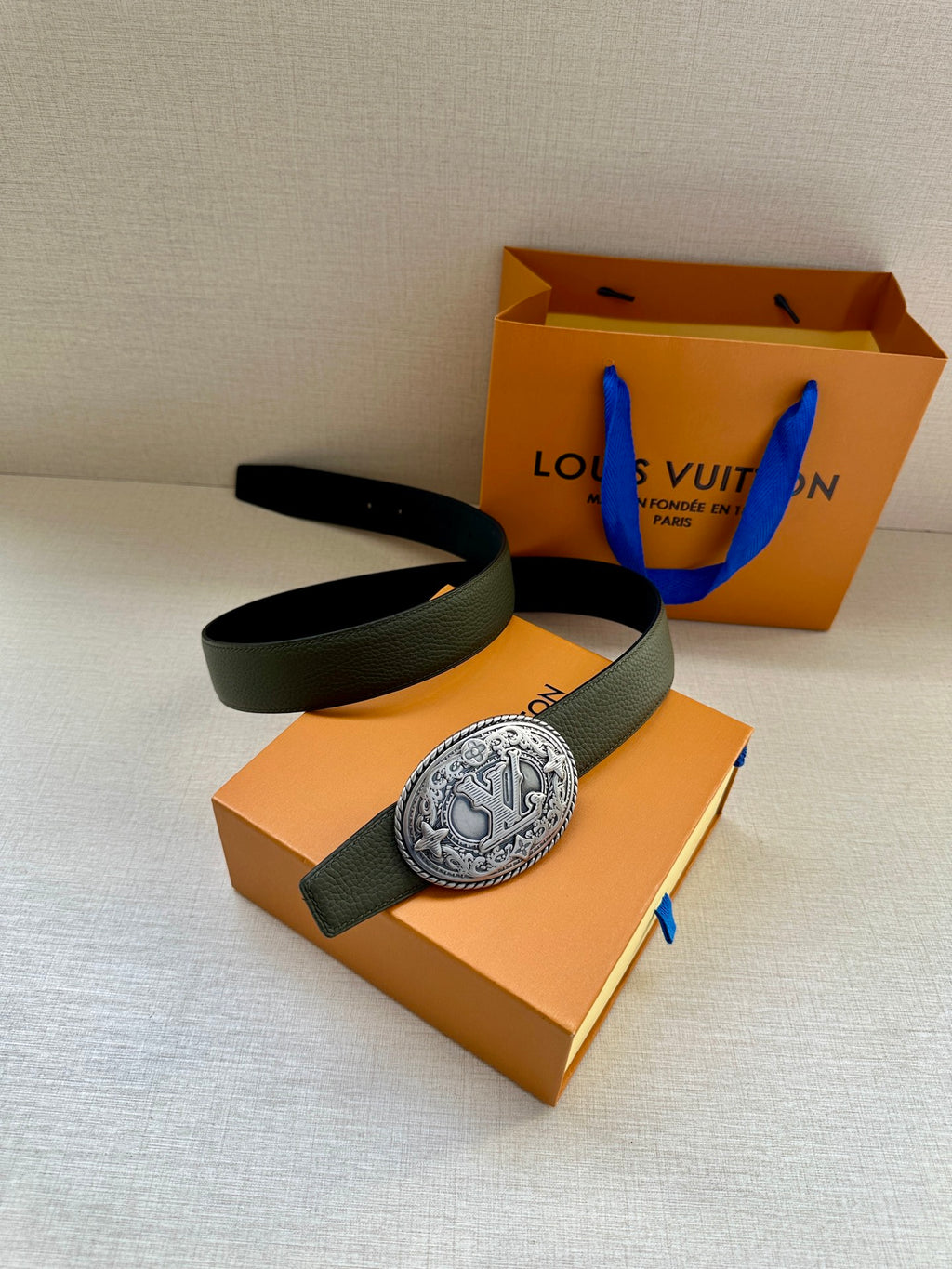 Dark green leather belt with large oval metal buckle, laid over gift box.