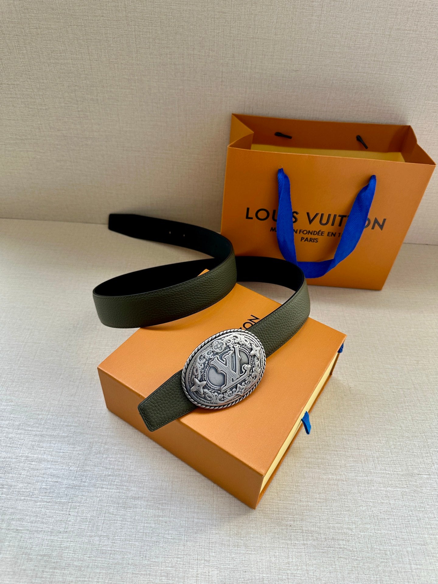Dark green leather belt with large oval metal buckle, laid over gift box.