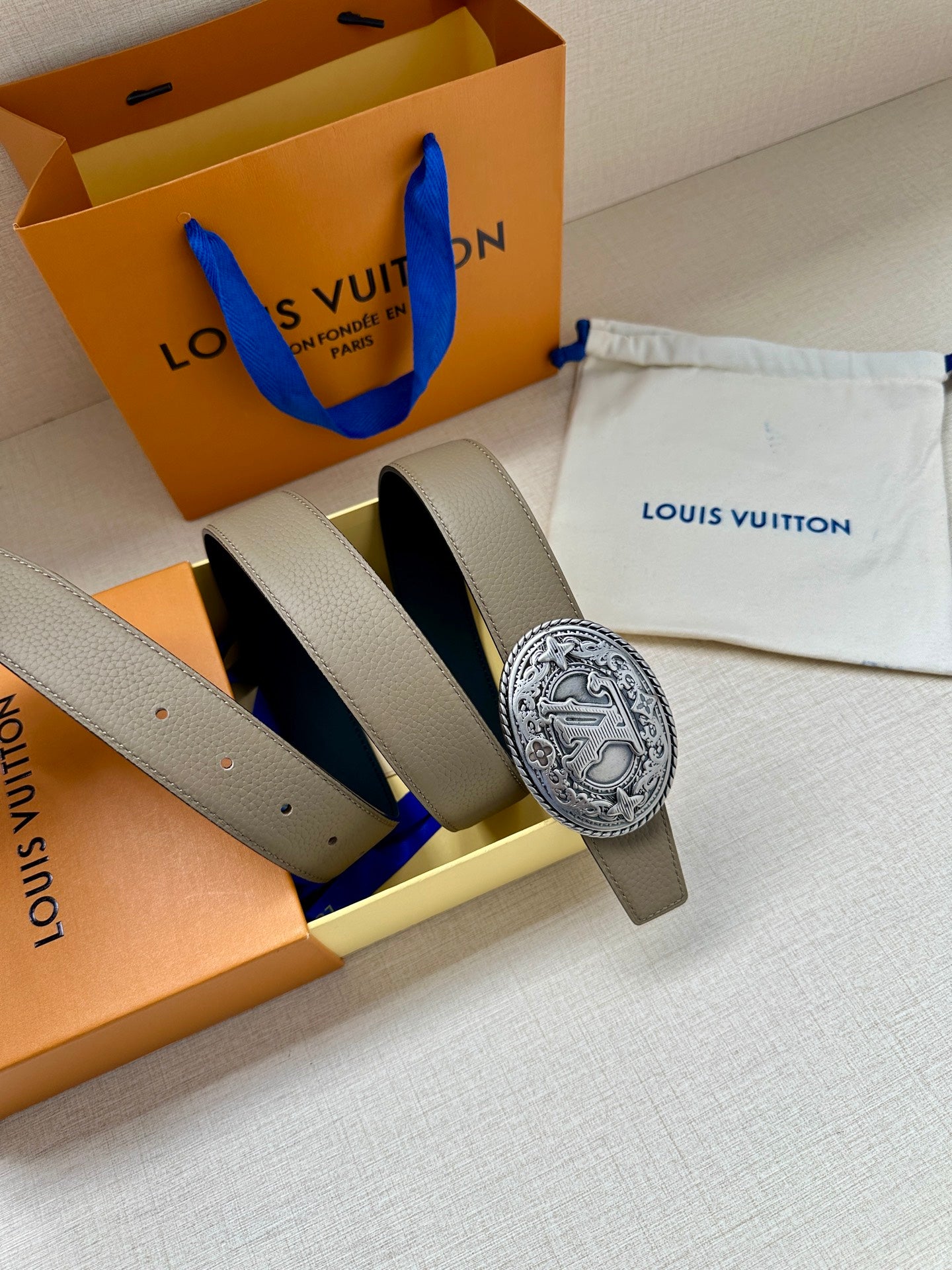 Louis Vuitton belt with silver-toned oval logo buckle on beige strap, angled view