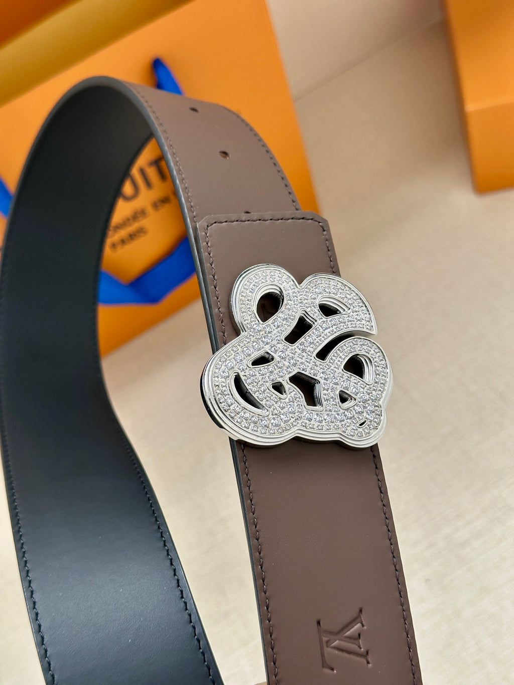 Close-up of brown belt strap with silver monogram buckle and stitched edge detail.
