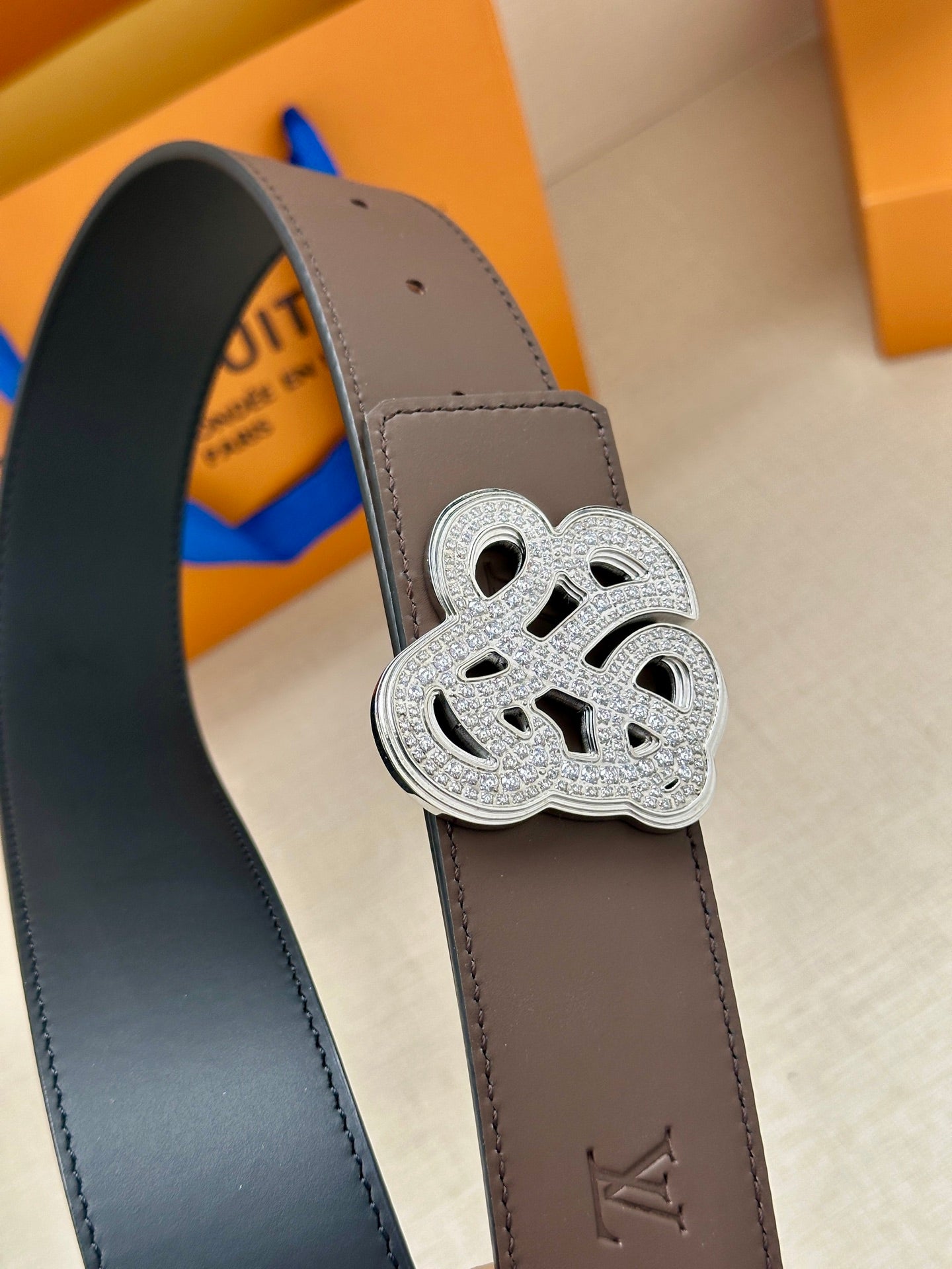 Close-up of brown belt strap with silver monogram buckle and stitched edge detail.