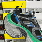 Black sneaker with gray side panels, blue logo stripe, green sole edge, and gray tread