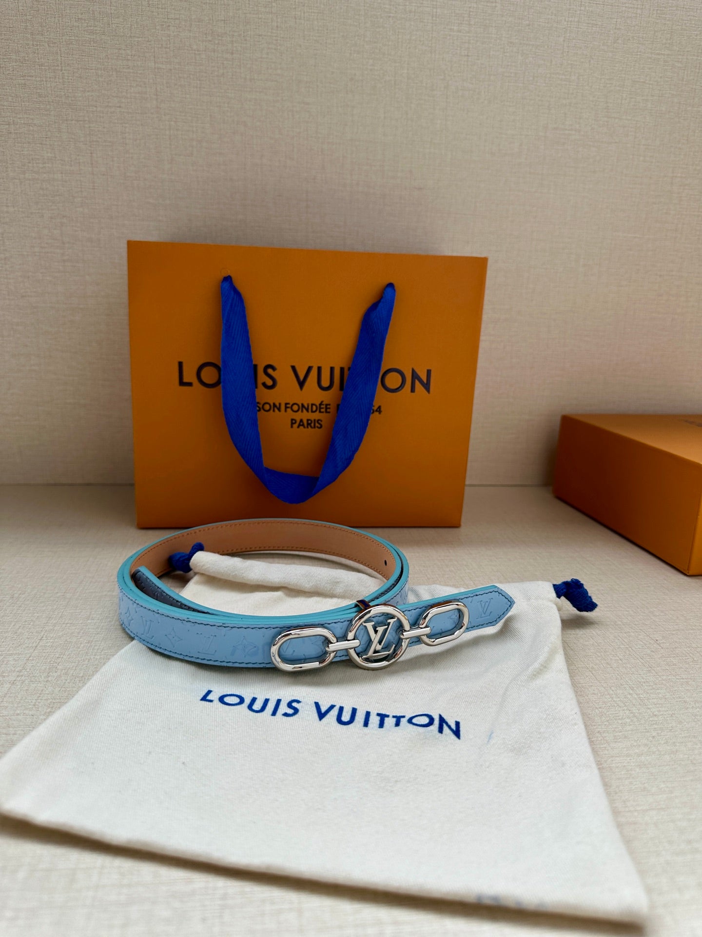 Light blue leather belt with gold LV buckle on a white dust bag.