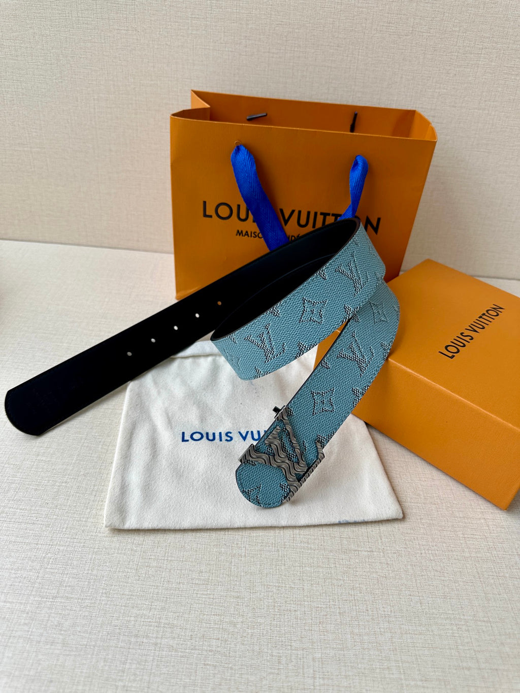 Blue monogram belt with gold buckle and black underside, next to dust bag.