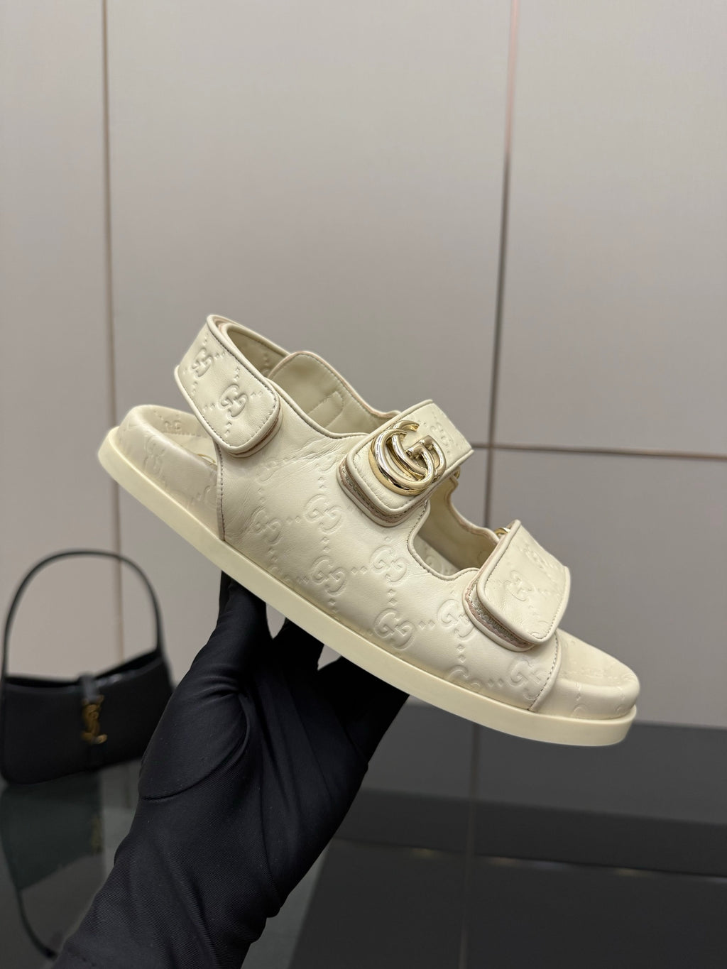 Cream slide sandals held in hand; metal buckle and embossed logo strap