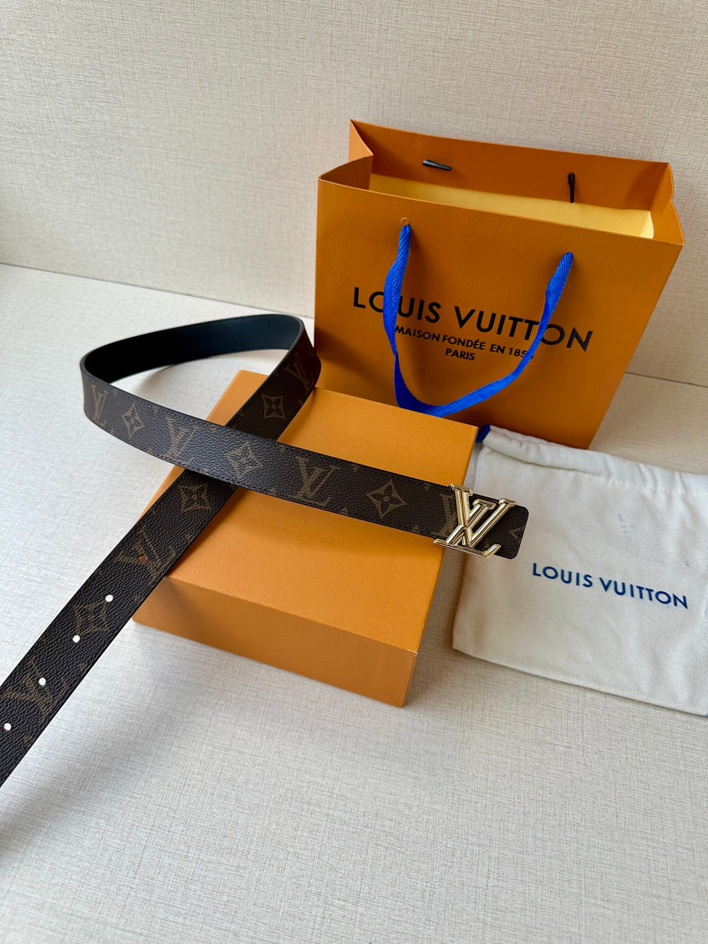 Brown monogram leather belt with gold-tone buckle and shopping bag beside dust cloth