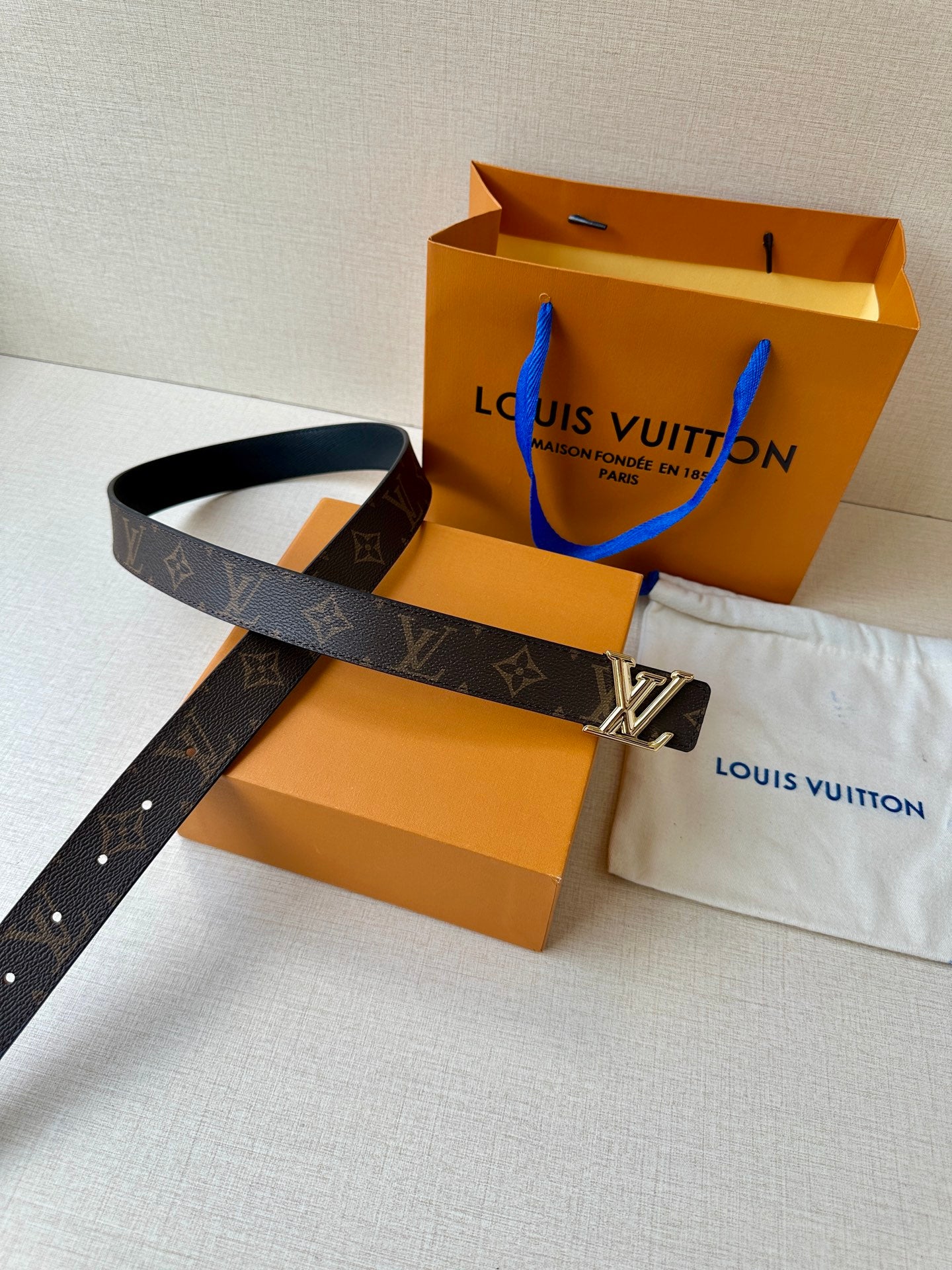 Brown monogram leather belt with gold-tone buckle and shopping bag beside dust cloth