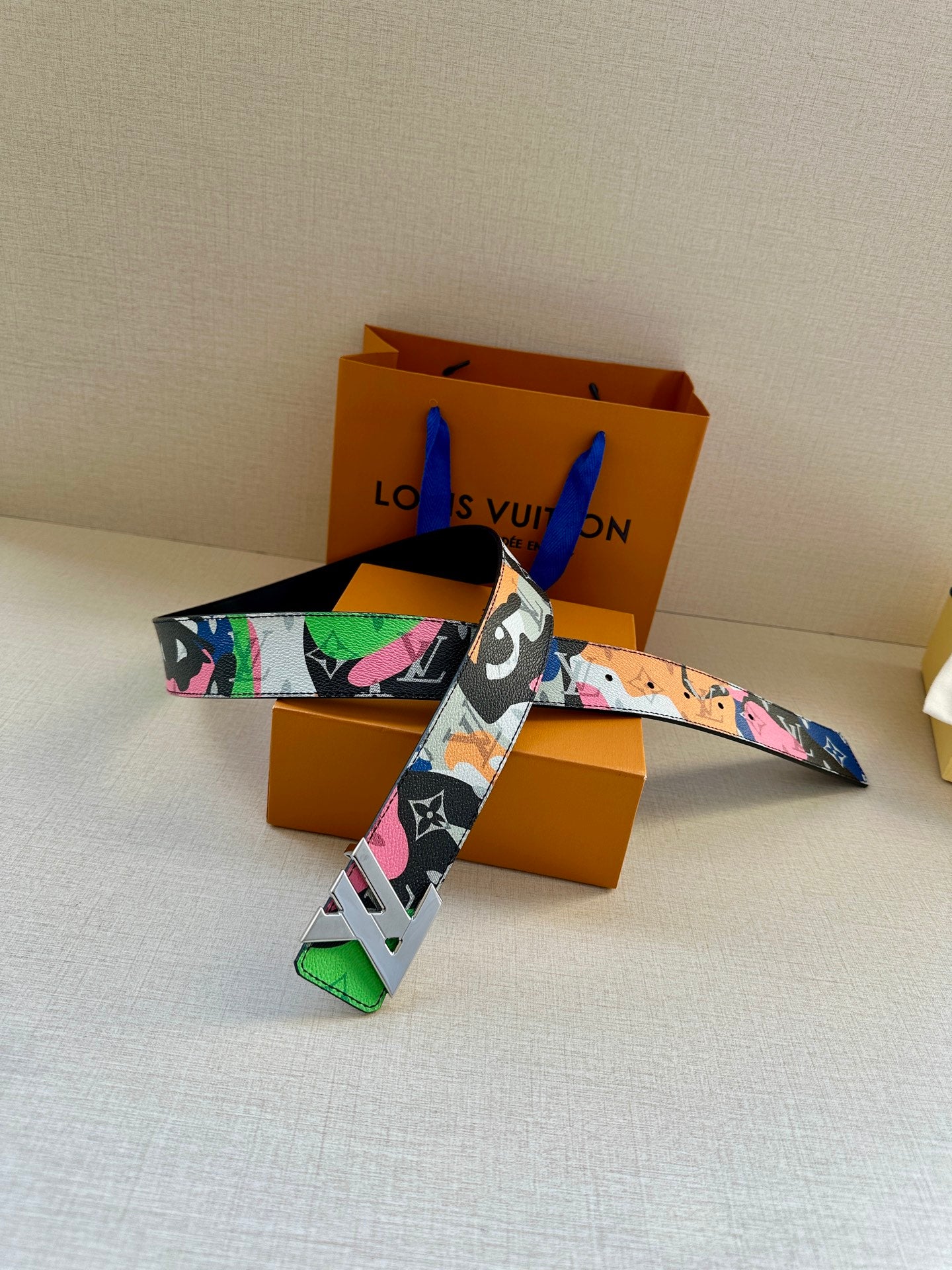 Designer belt laid flat on white fabric; multicolor patterned strap and gold buckle