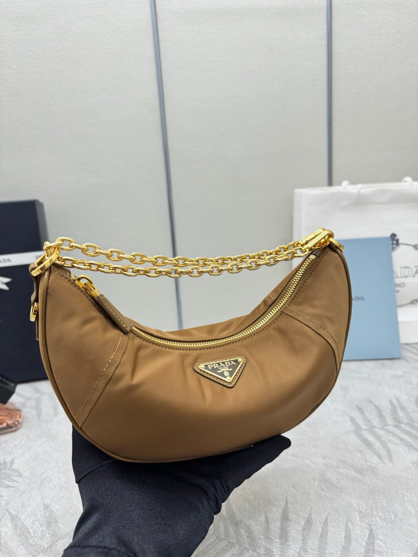 Prada tan leather hobo bag with gold chain strap, front crest logo, and zip opening.