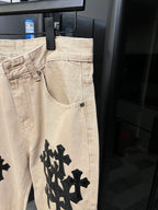 Beige denim pants with black cross patch on lower front leg and pocket