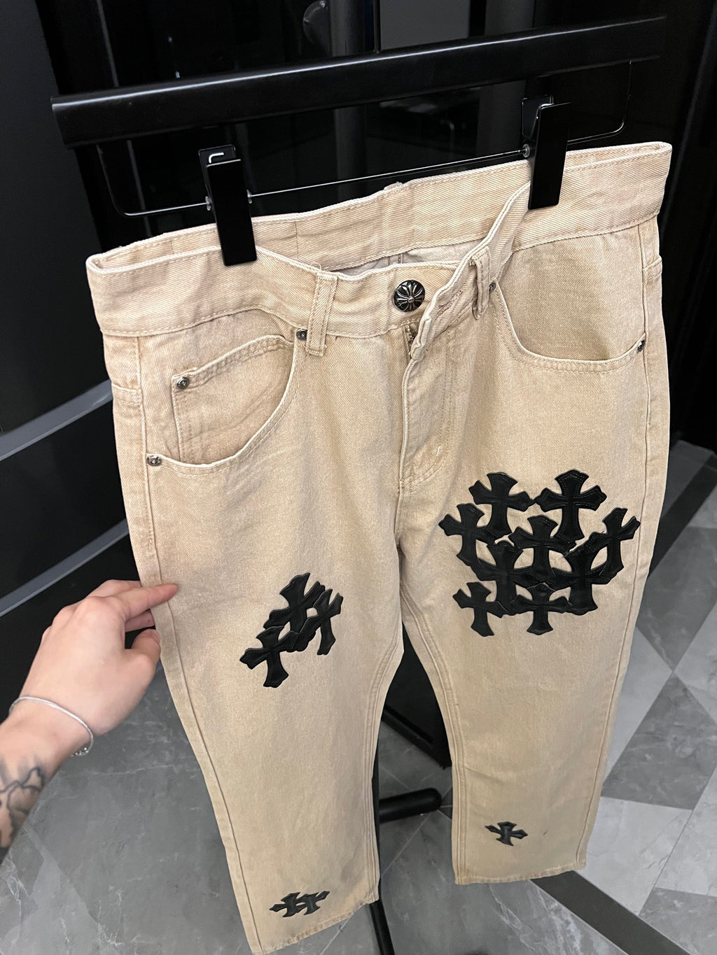 Beige denim pants with black cross patch on right leg and five-pocket styling