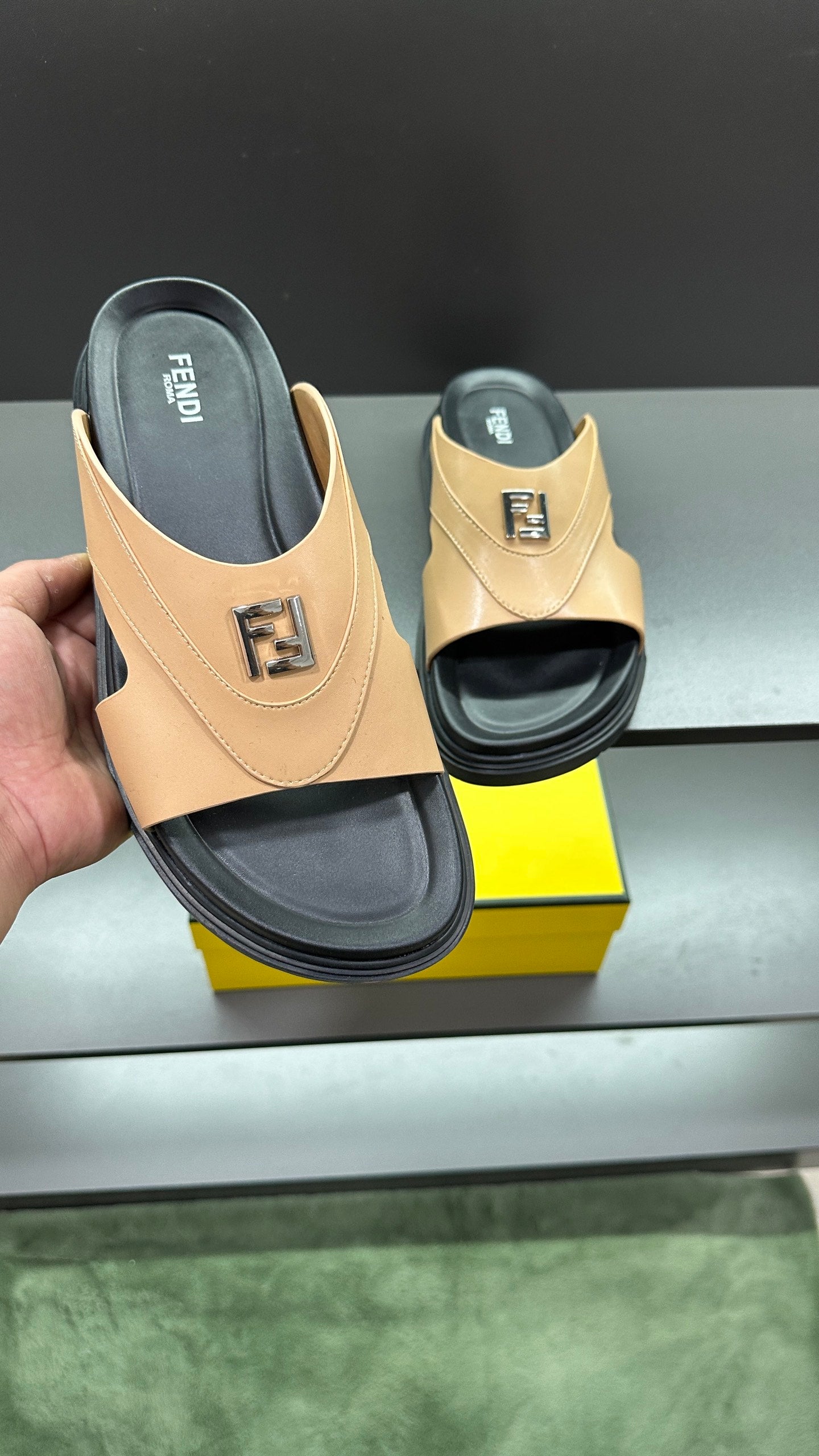 Hand holding Fendi beige slide sandals; black footbed and F logo on upper