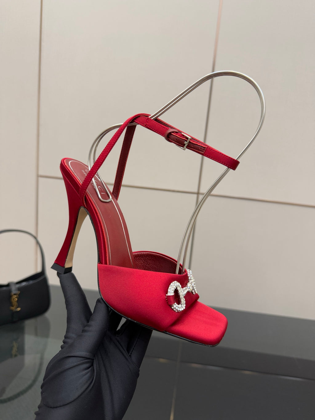Red heeled sandal with clear ankle strap and silver buckle detail, held in black glove